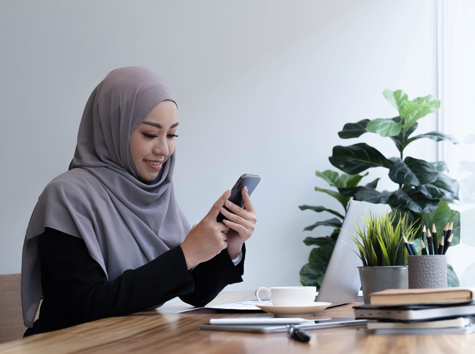 Positive beautiful young muslim woman in purple headscarf using brand new mobile phone, looking at copy space and smiling, using newest mobile application for business, home interior.